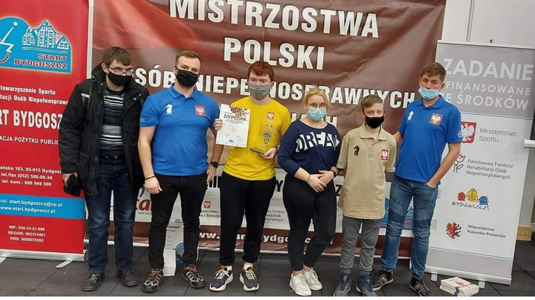 Grupa osób w maseczkach z dyplomem na tle banera mistrzostw polski, klub szachowy KSN Łuczniczka Bydgoszcz