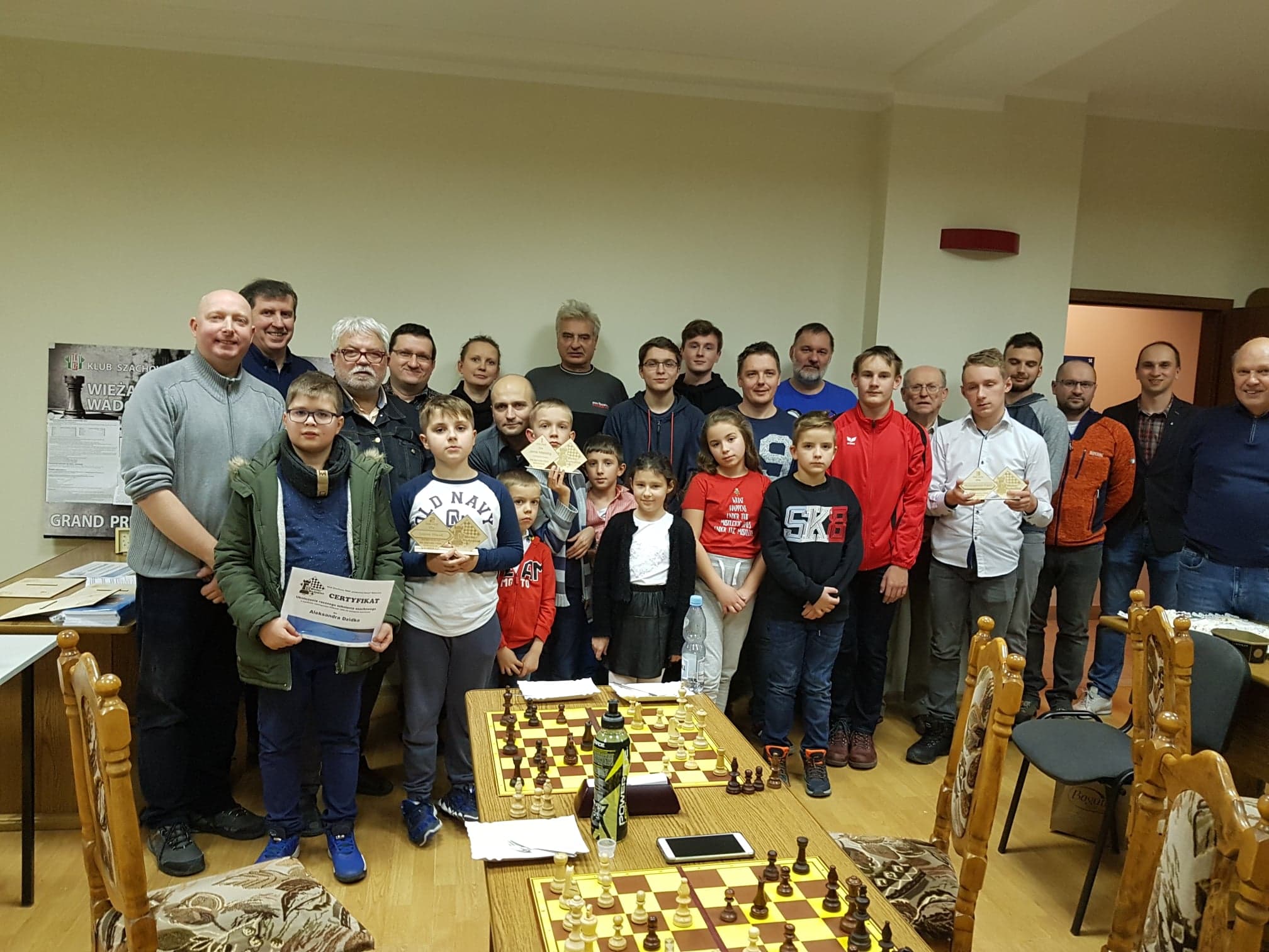 Duża grupa osób z dyplomami przy szachownicach, klub szachowy TKKF Leskowiec - Wieża Wadowice