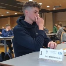Młody zawodnik siedzi przy stoliku turniejowym z tabliczką klubu, trener szachów Jan Golenia z Wieliczki